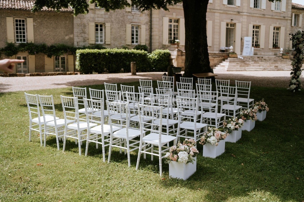 Chaises blanches alignées en extérieur pour une cérémonie, entourées de fleurs, devant un bâtiment ancien.