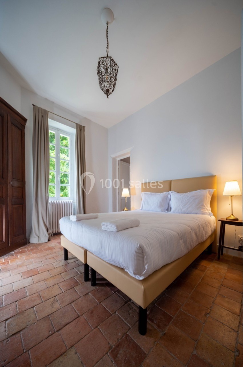 Chambre lumineuse avec lit double, sol en terre cuite, armoire en bois, fenêtre donnant sur un espace verdoyant.
