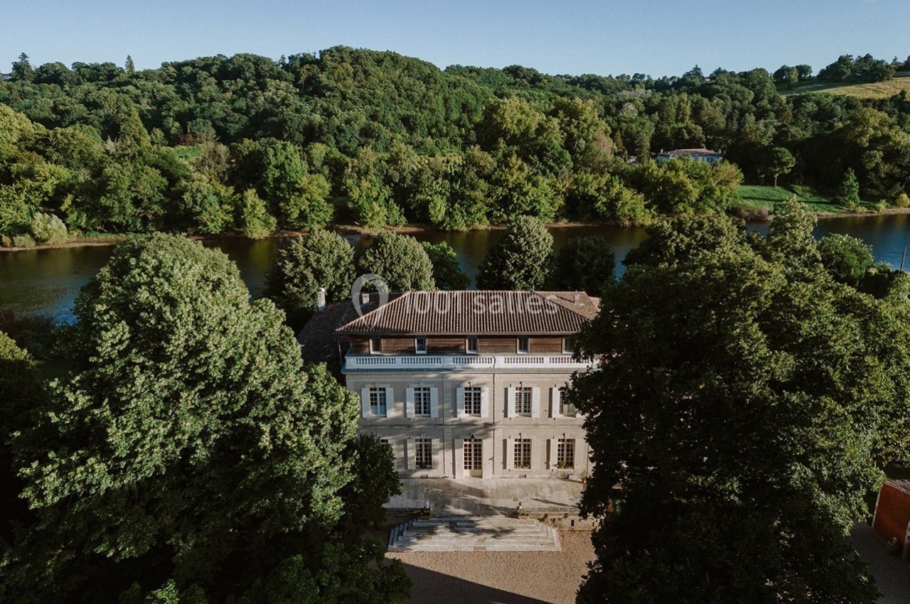 Vue aérienne d'un manoir entouré d'arbres, situé près d'une rivière sous un ciel dégagé.