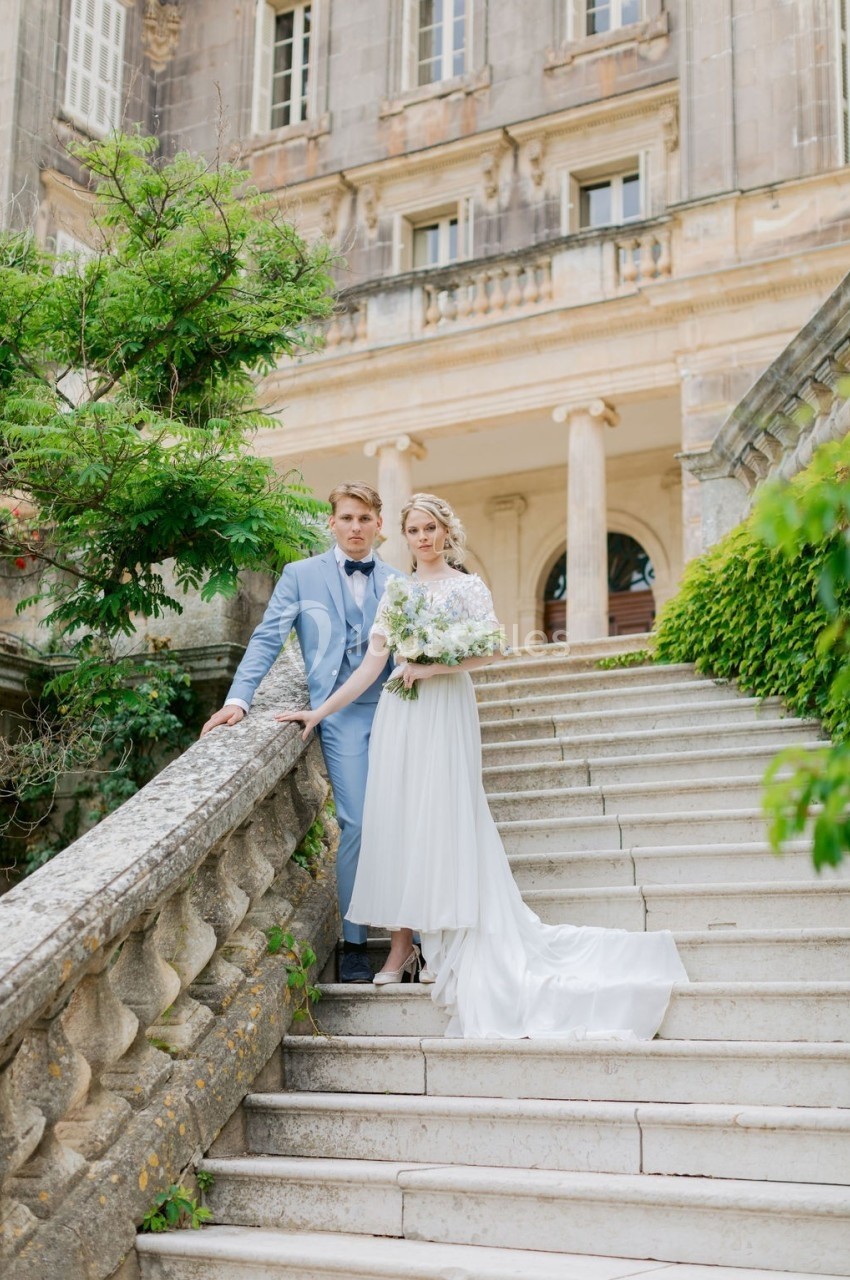 Un couple en tenue de mariage pose sur un escalier en pierre devant un bâtiment ancien entouré de verdure.