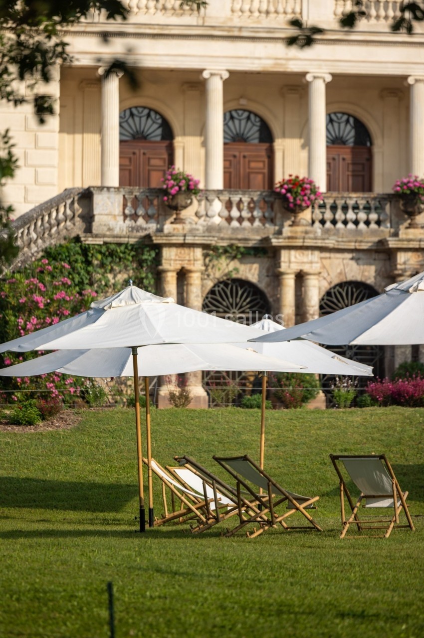 Chaises longues et parasols blancs disposés sur une pelouse devant un bâtiment ancien avec colonnes et balustrades.