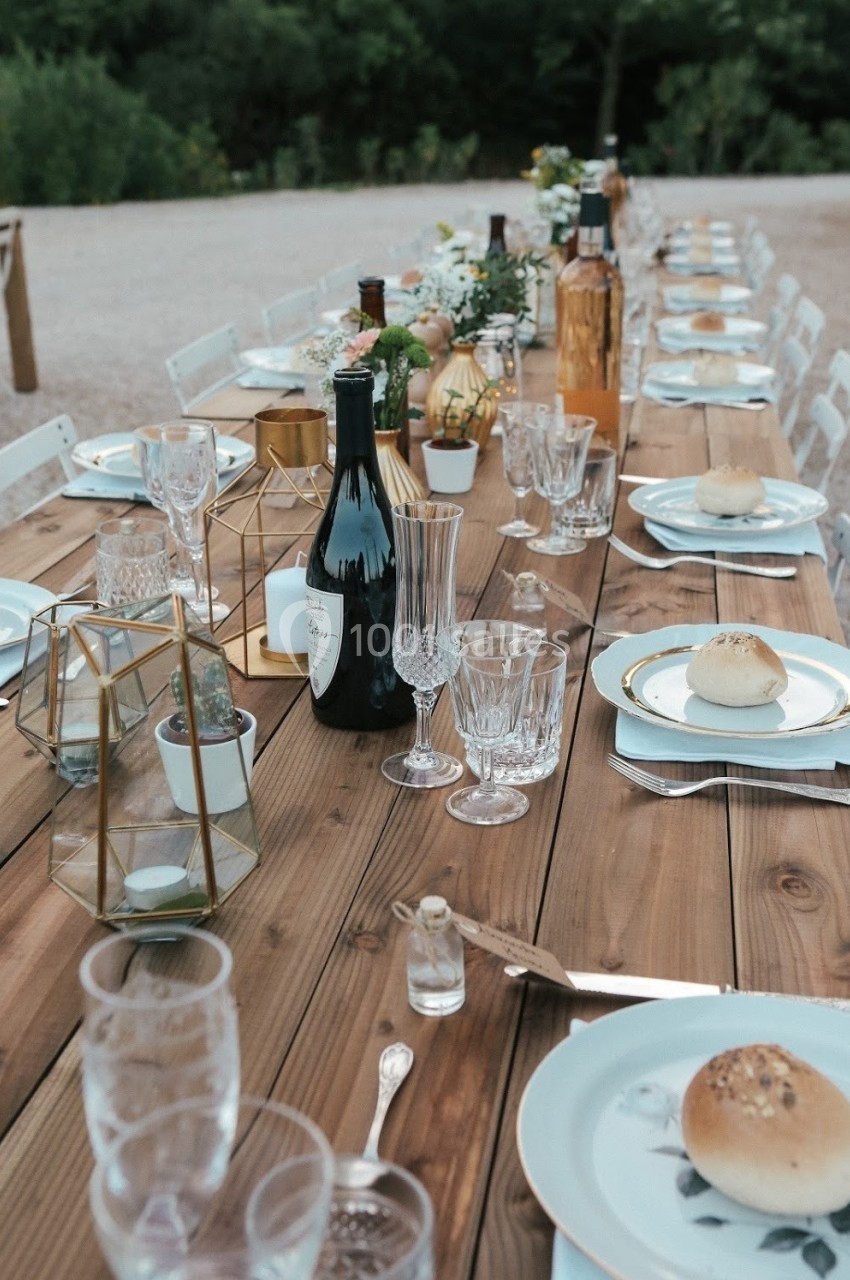 Table en bois dressée pour un repas, avec assiettes, verres, bouteilles, pains et décorations florales.