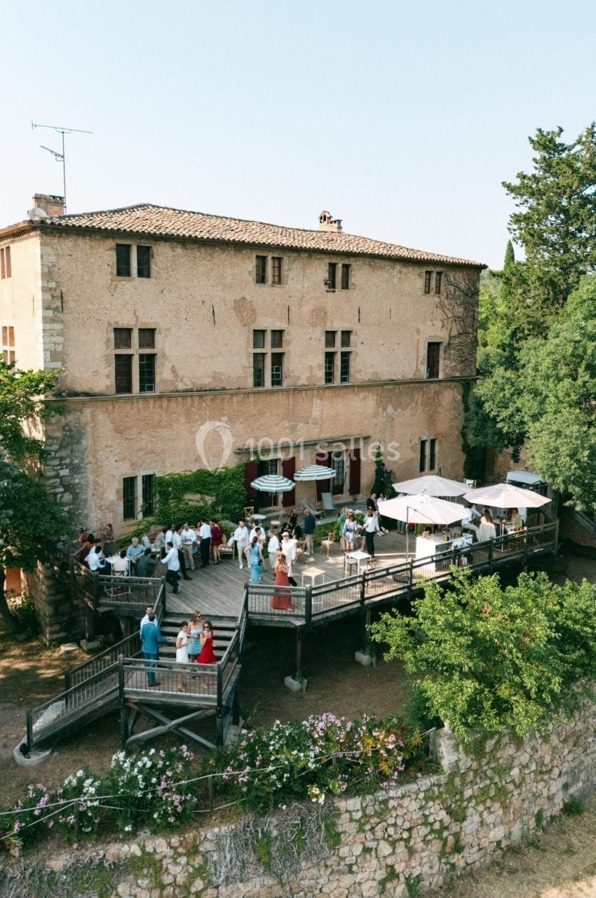 Groupe de personnes rassemblées sur une terrasse en bois devant un grand bâtiment en pierre entouré de verdure.