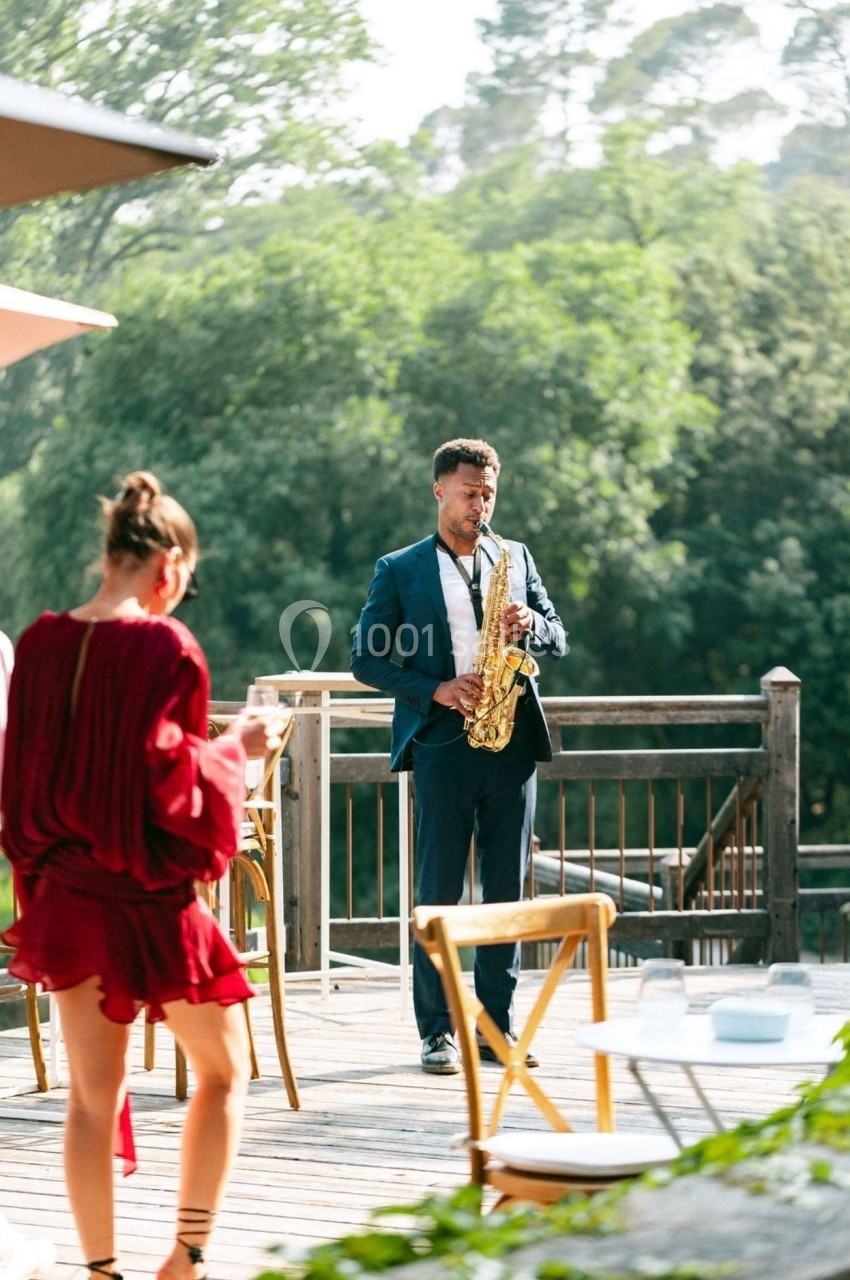 Un musicien en costume joue du saxophone sur une terrasse en bois entourée de verdure, avec des invités en arrière-plan.