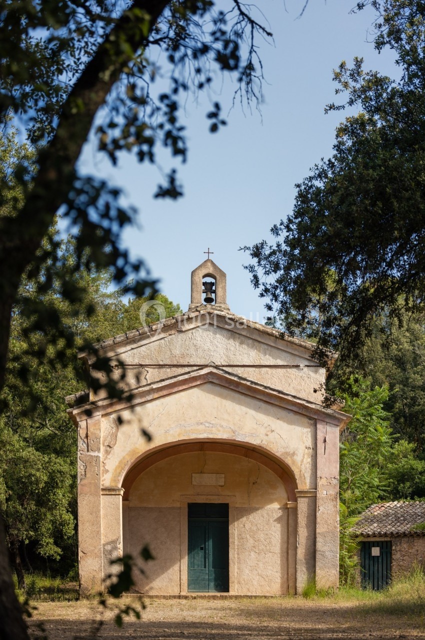 Petite chapelle ancienne entourée de végétation, avec une façade simple et un clocher surmonté d'une croix.