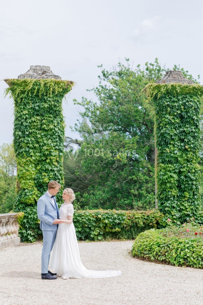 Un couple en tenue de mariage se tient debout dans un jardin avec des colonnes couvertes de lierre en arrière-plan.