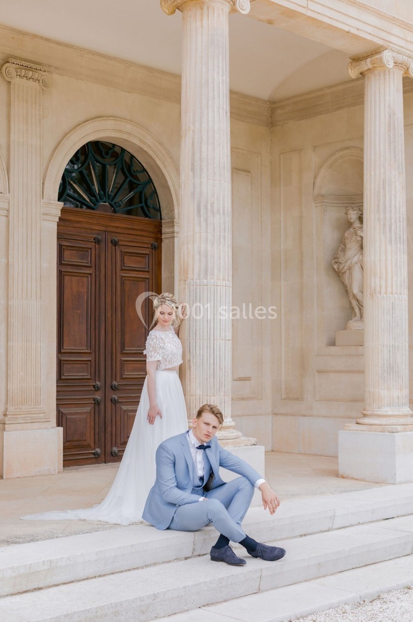 Un couple en tenue de mariage posé devant un bâtiment classique avec colonnes et porte en bois sculpté.