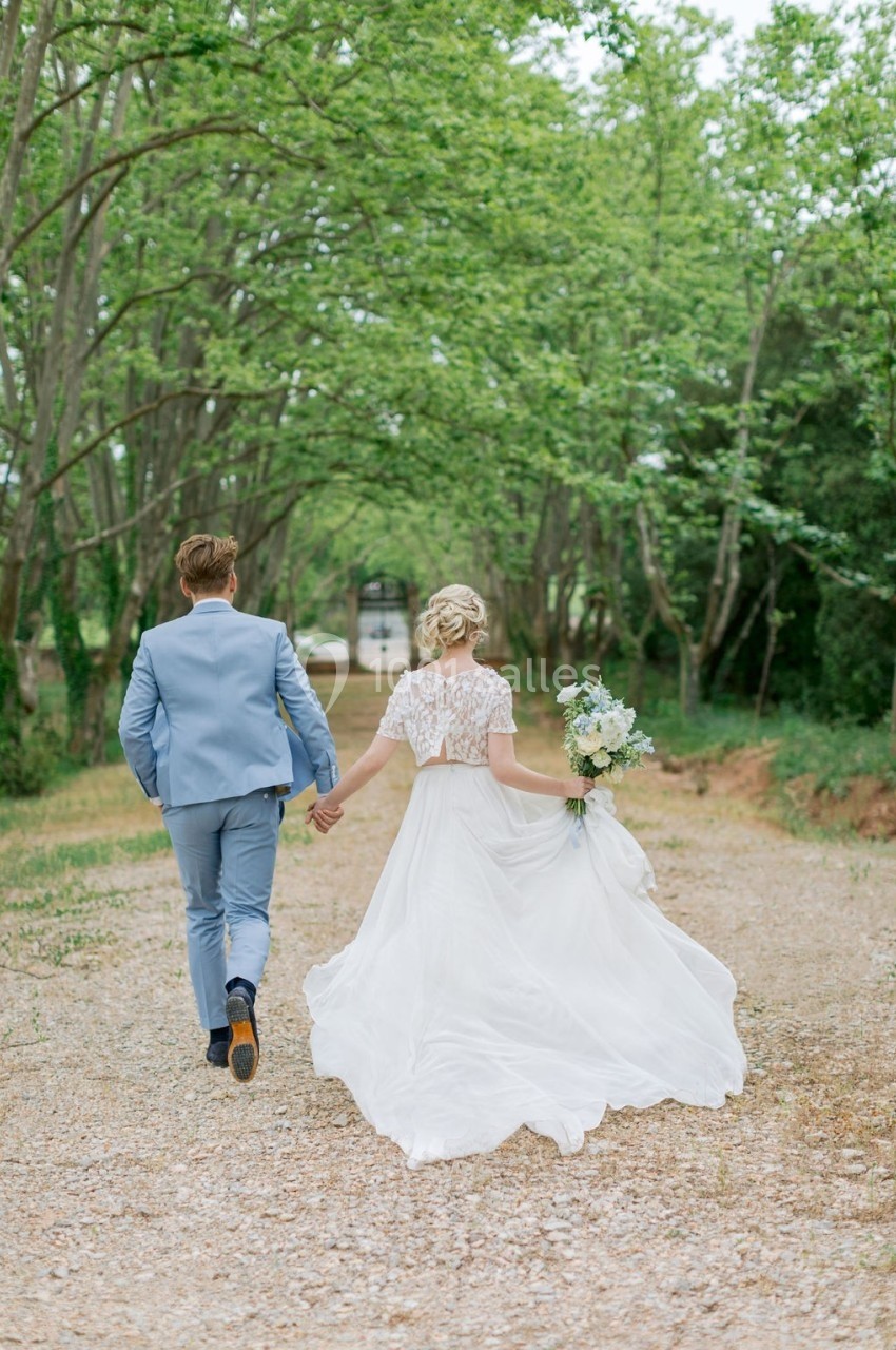 Un couple en tenue de mariage marche main dans la main sur un chemin bordé d'arbres.