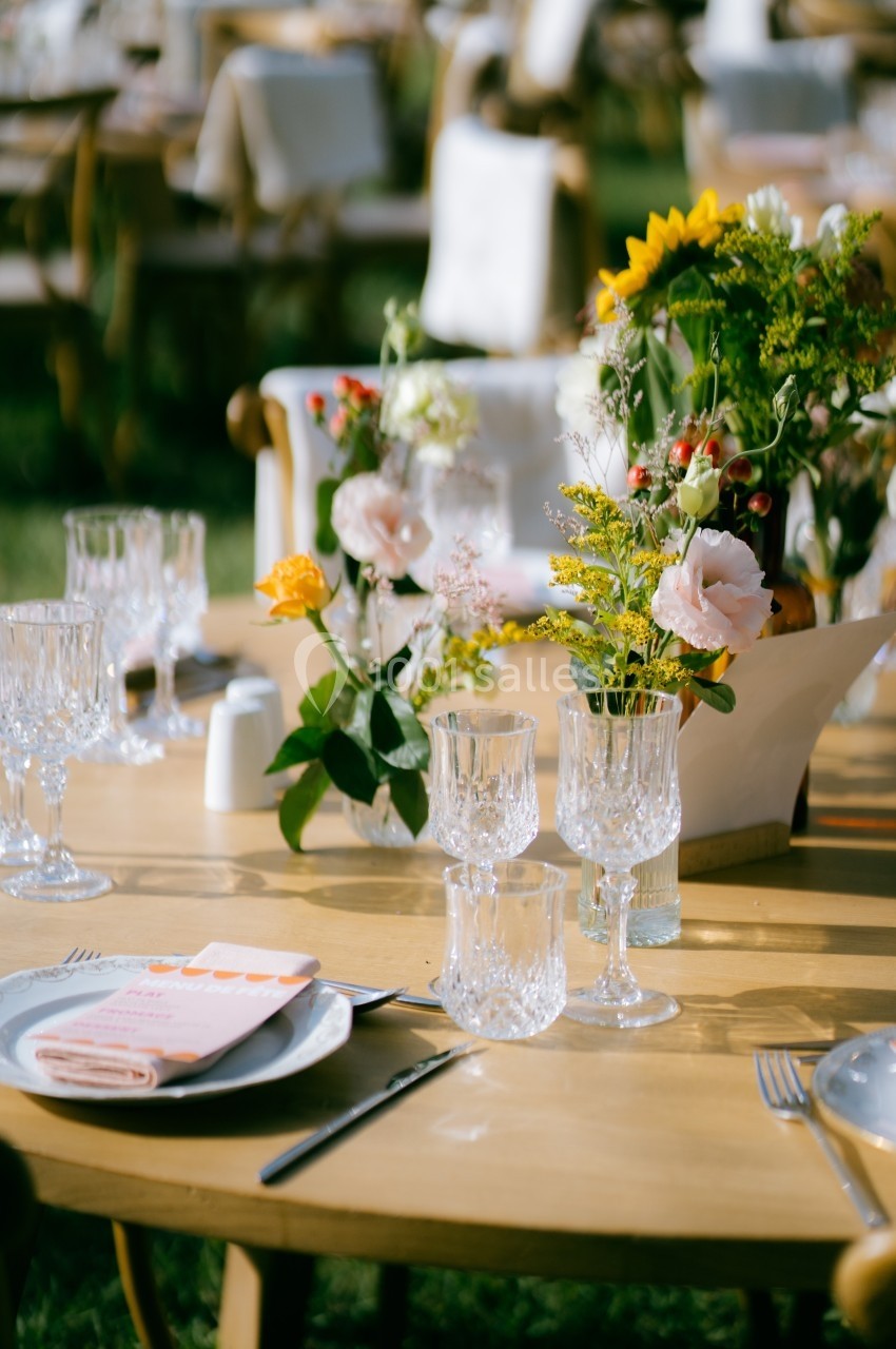 Table en bois décorée avec des fleurs colorées, des verres en cristal et des serviettes, dans un cadre extérieur.