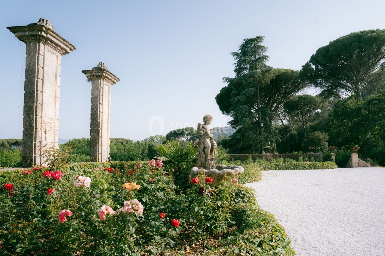 Jardin avec des colonnes antiques, une statue et des rosiers fleuris, entouré de végétation et d'arbres.