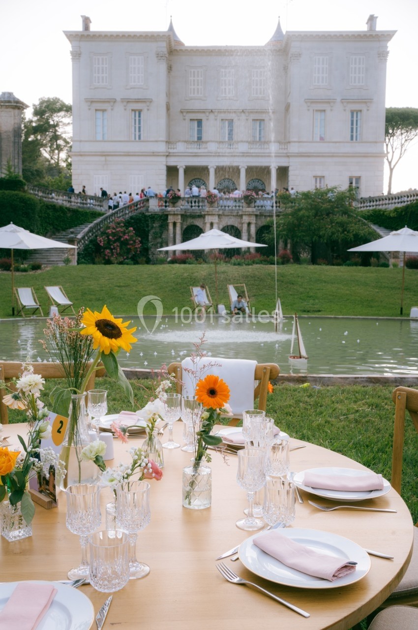 Table élégamment dressée avec des fleurs et des verres, devant un jardin avec fontaine et un bâtiment historique en arrière…