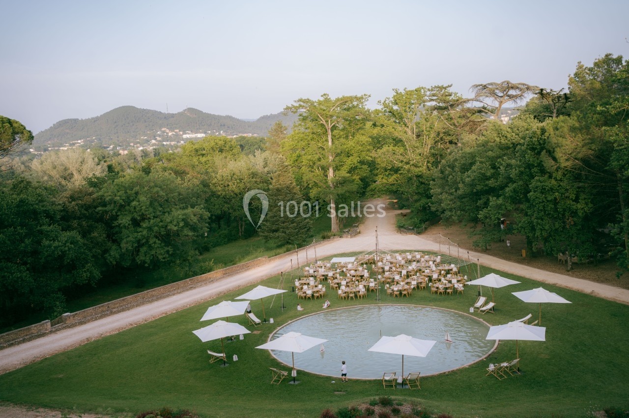Vue aérienne d'un jardin avec une petite étendue d'eau entourée de tables, chaises et parasols, dans un cadre verdoyant.