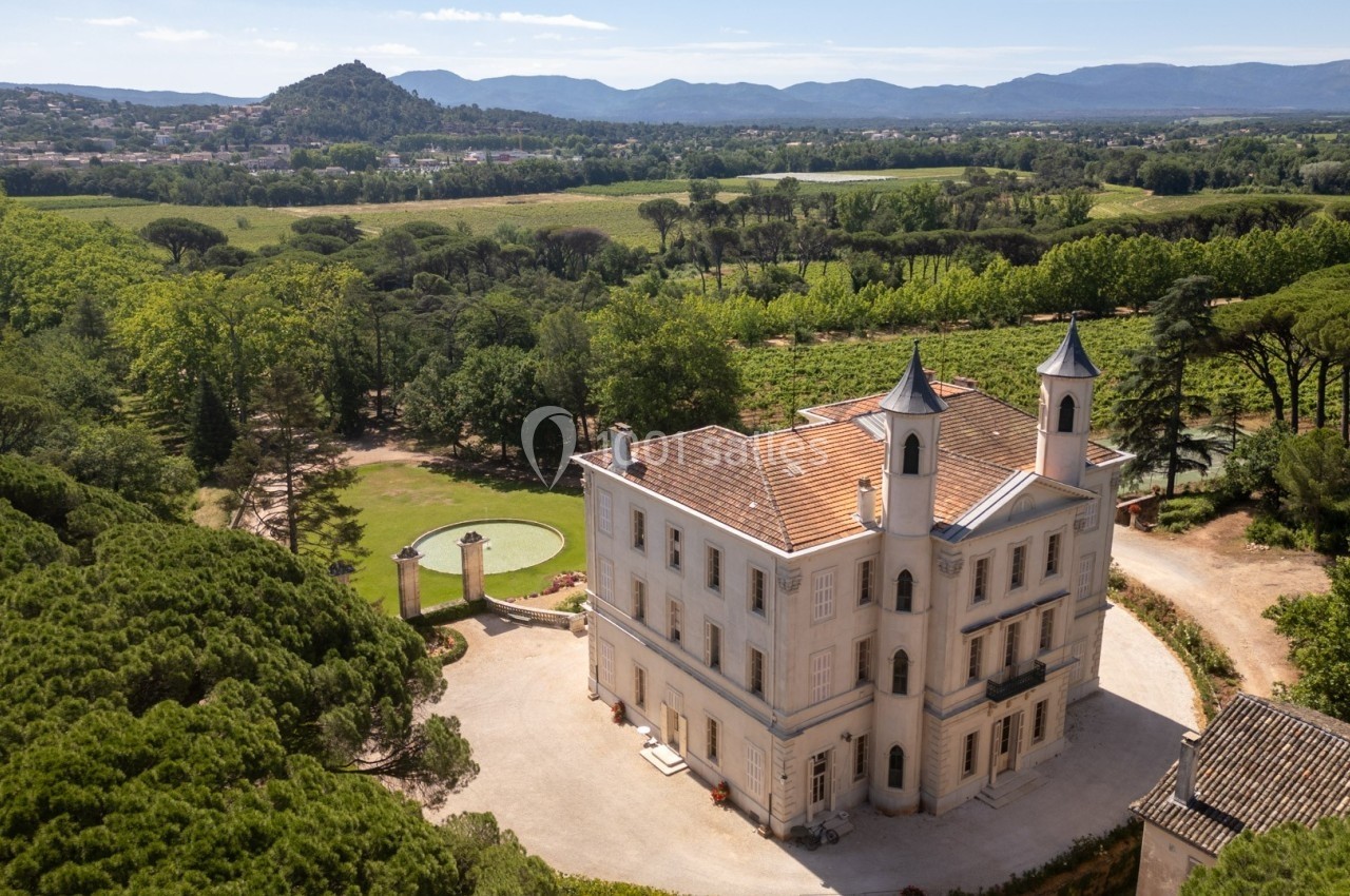 Vue aérienne d'un château entouré de vignes et de forêts, avec des collines en arrière-plan sous un ciel dégagé.