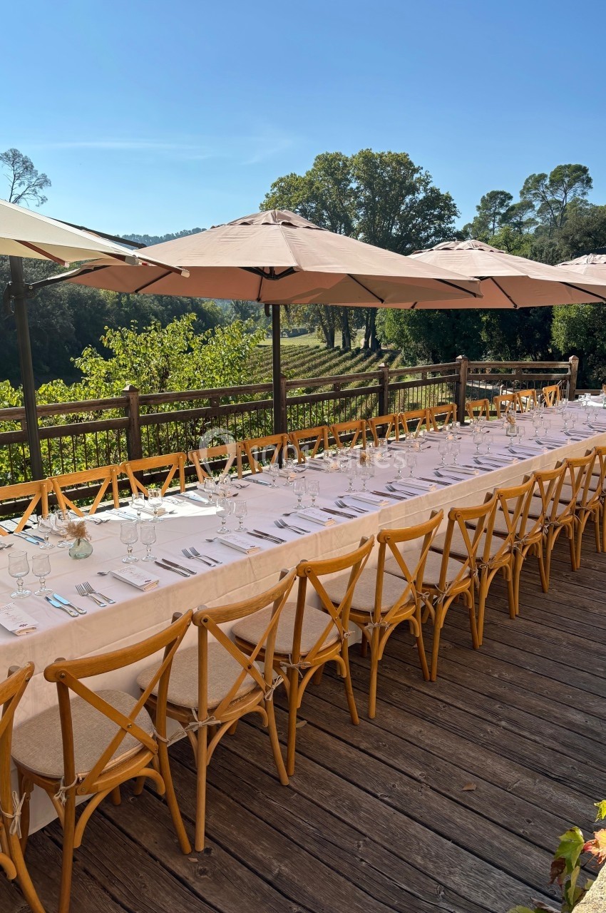 Grande table dressée avec nappes blanches, chaises en bois et parasols, sur une terrasse surplombant un paysage verdoyant.