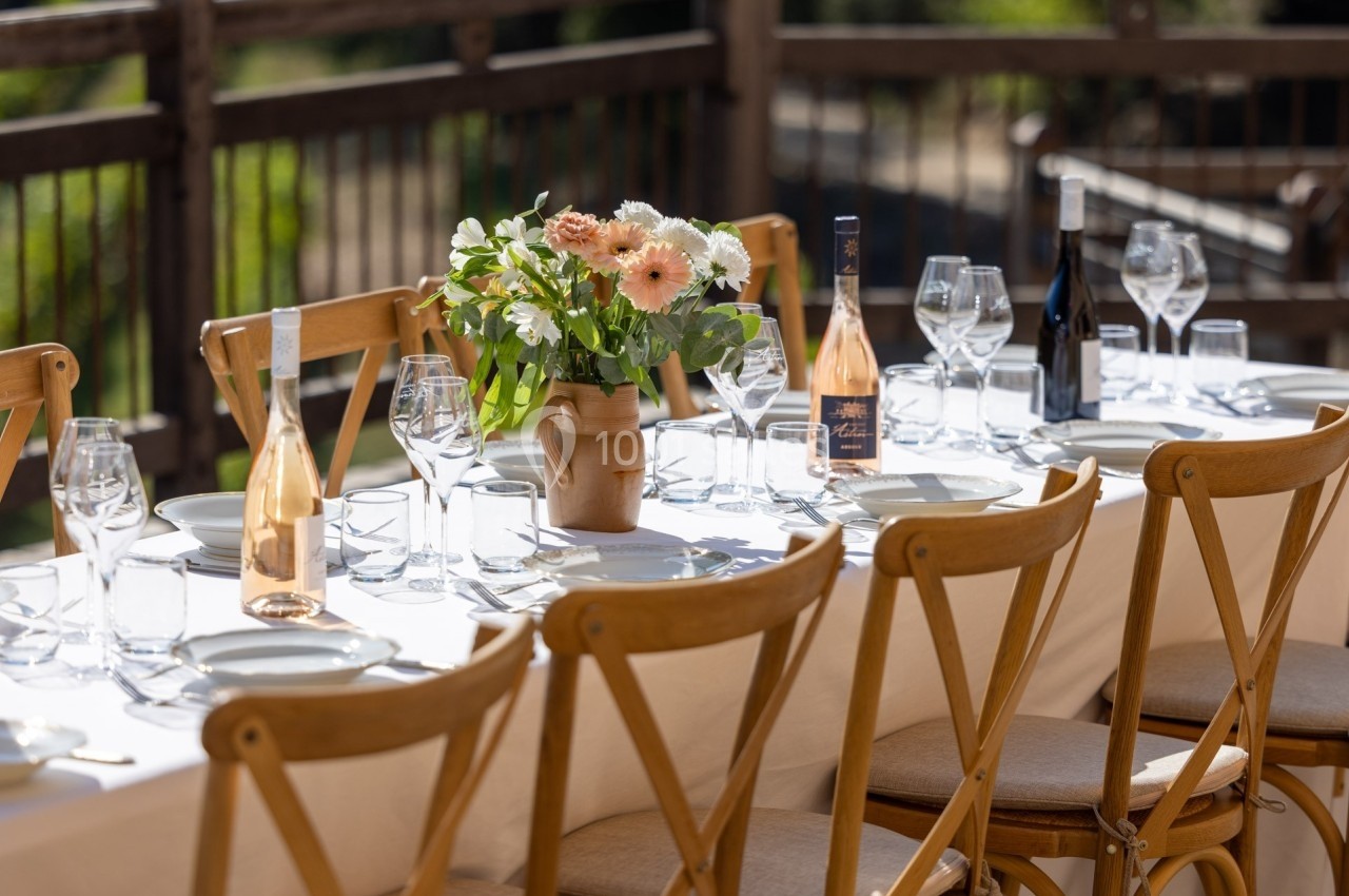 Table dressée en extérieur avec des chaises en bois, des verres, des bouteilles de vin et un bouquet de fleurs au centre.
