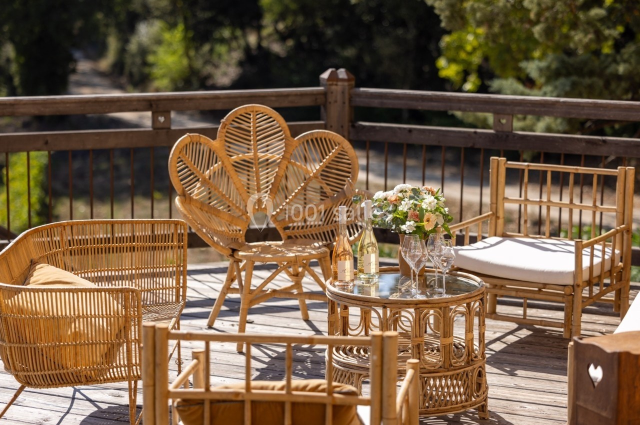 Mobilier en rotin disposé sur une terrasse en bois, avec table basse, fleurs et verres à vin.