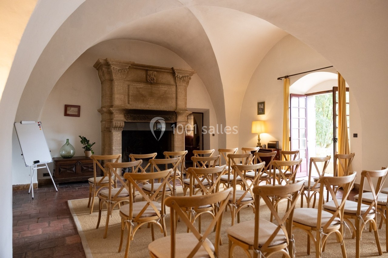 Salle voûtée aménagée avec des chaises en bois face à une cheminée en pierre, éclairée par une lumière naturelle.