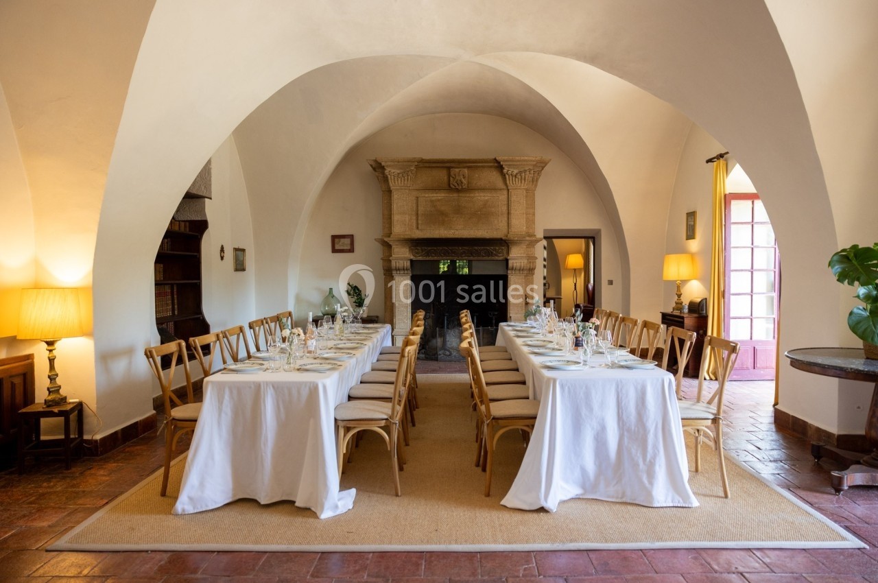 Salle voûtée avec deux longues tables dressées pour un repas, entourées de chaises en bois et éclairées par des lampes et la…