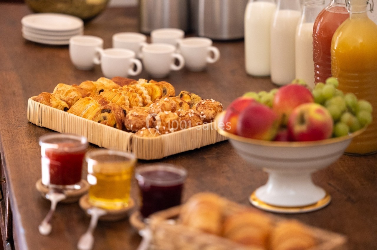 Plateau de viennoiseries entouré de confitures, fruits, jus et tasses sur une table en bois.