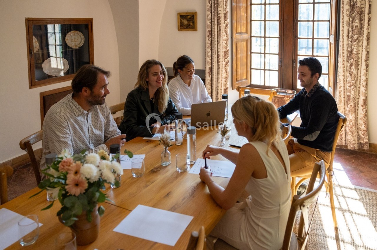Un groupe de personnes discute autour d'une table en bois dans une pièce lumineuse avec des fenêtres et des fleurs.