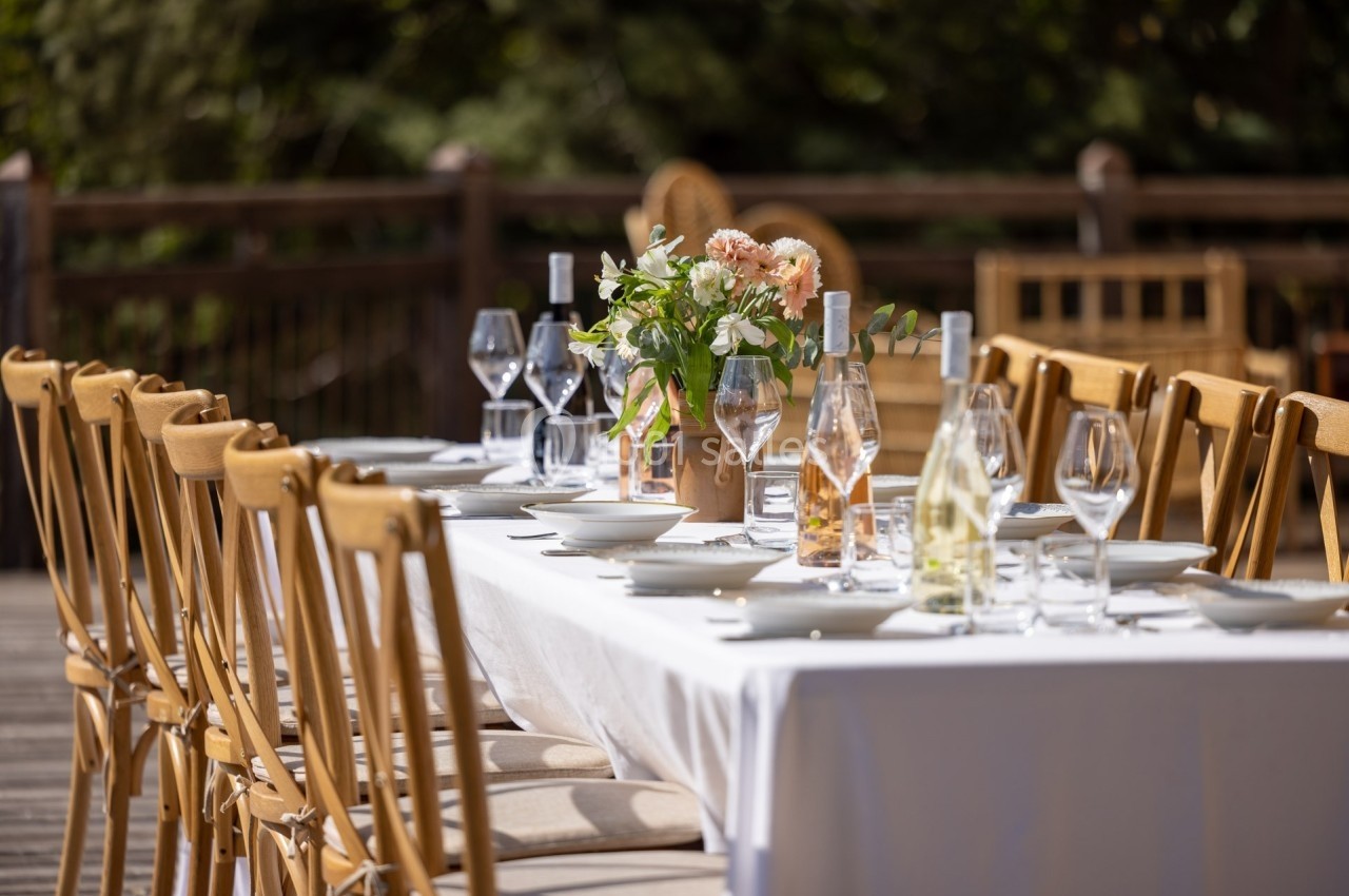 Table dressée en extérieur avec nappes blanches, vaisselle, verres et un bouquet de fleurs au centre.