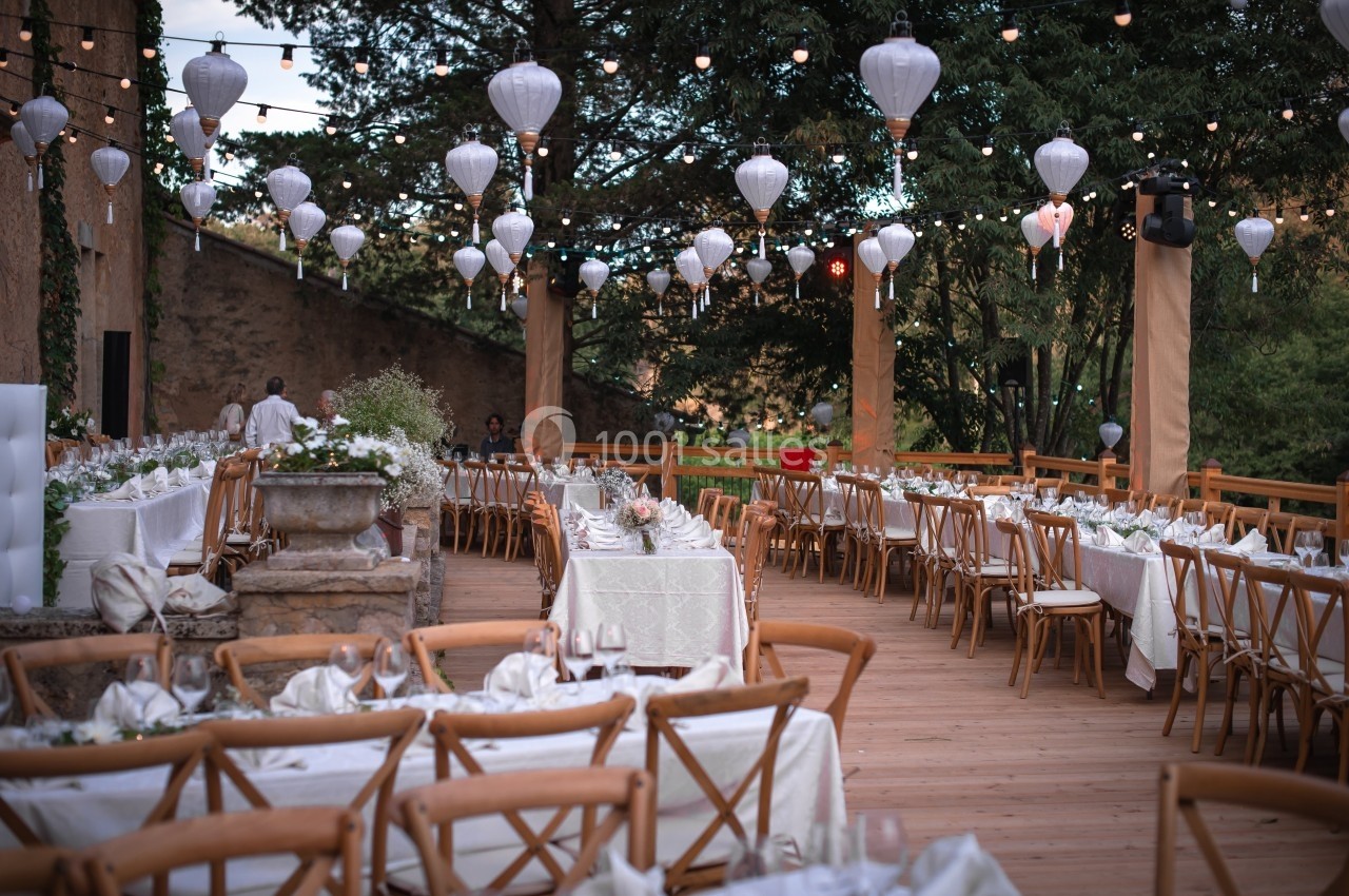 Terrasse décorée pour un événement, avec tables dressées, chaises en bois et lanternes suspendues sous des guirlandes…