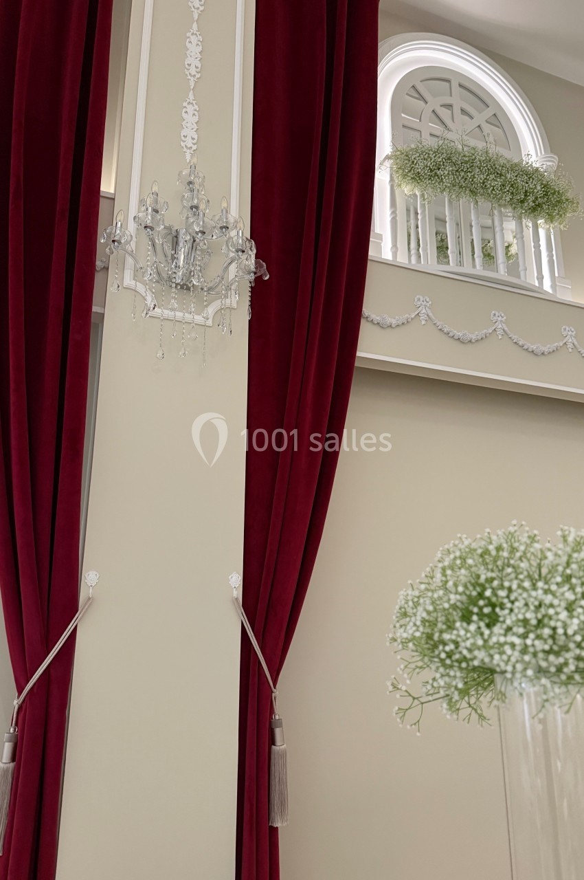 Rideaux rouges encadrant un mur beige décoré de moulures, avec un lustre en cristal et une balustrade ornée de verdure.