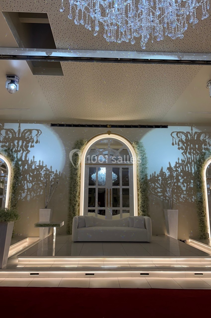 Salle élégante avec canapé blanc, arches lumineuses, décor végétal et lustre en cristal suspendu.