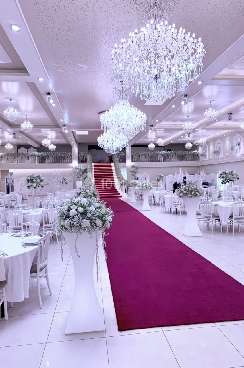 Salle de réception élégante avec tapis rouge, tables décorées de fleurs blanches et lustres suspendus.