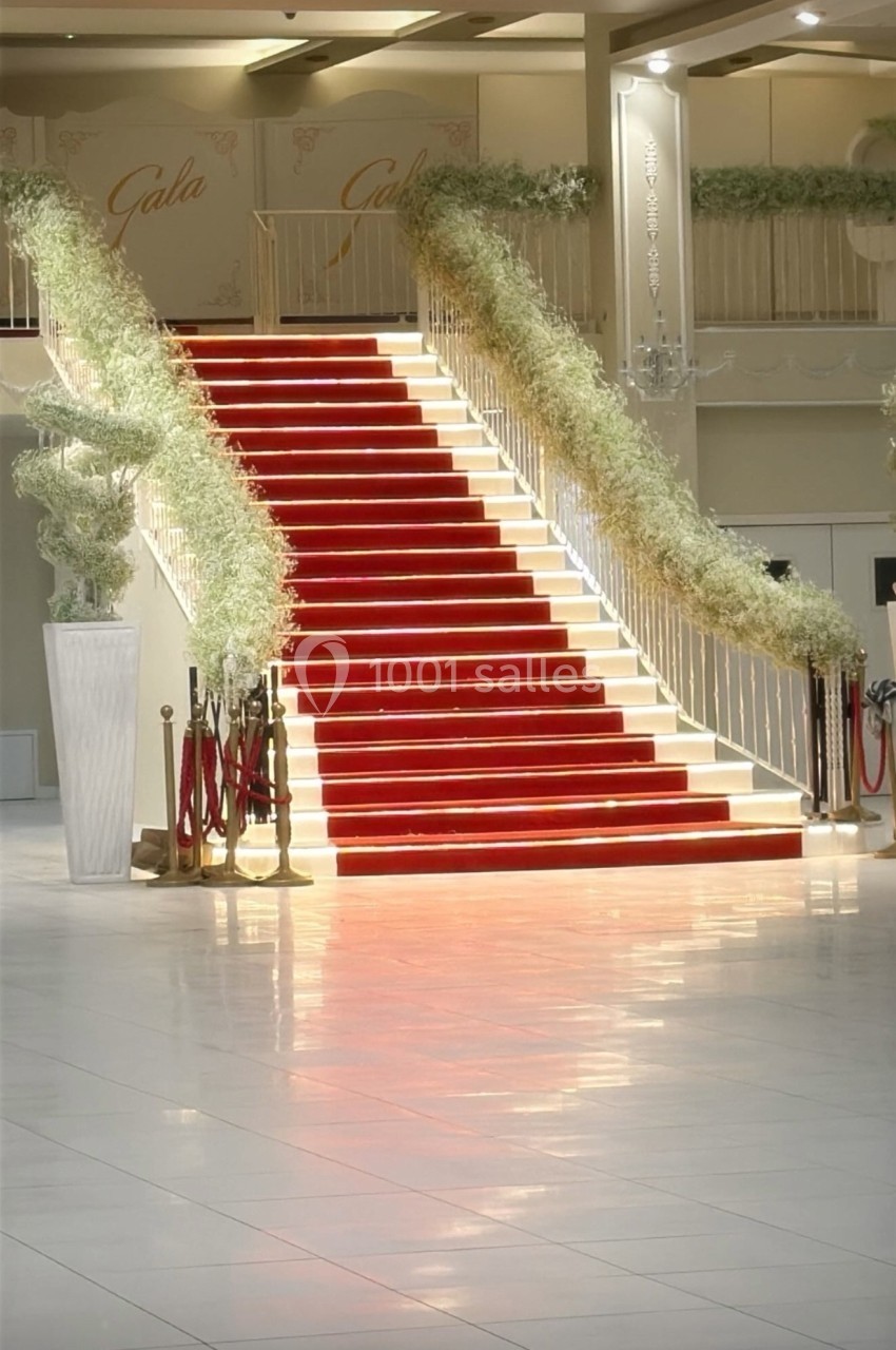 Escalier rouge bordé de rambardes dorées et décoré de plantes blanches, dans un espace intérieur lumineux.