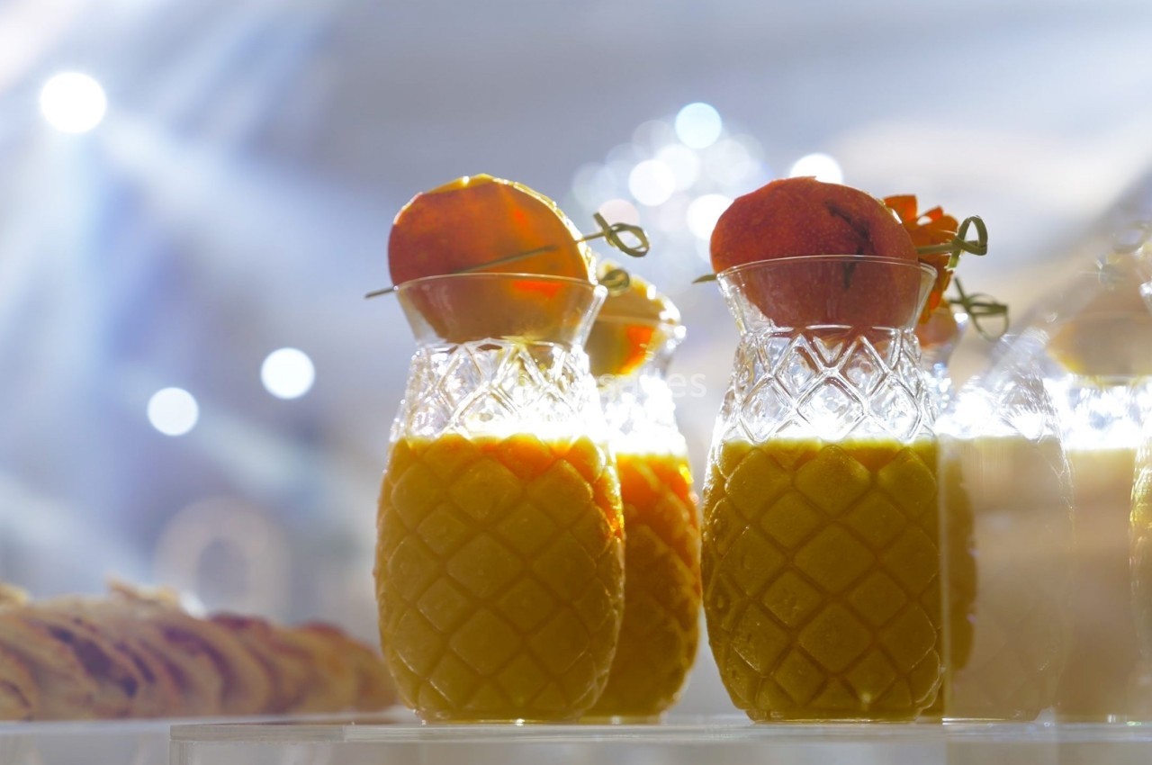 Verres en forme d'ananas remplis de jus orange, décorés de tranches de fruits, sur une table éclairée.