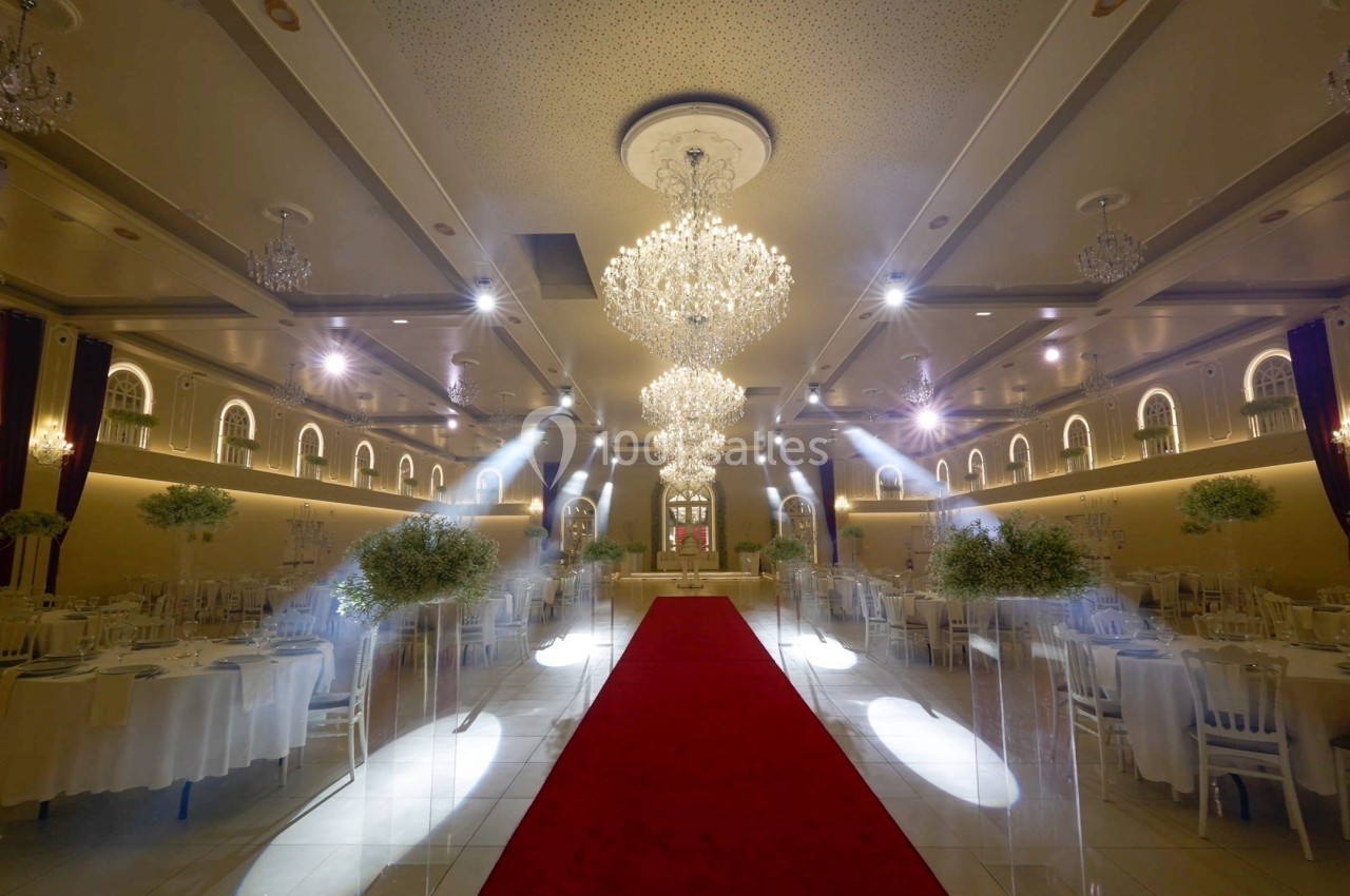 Salle de réception élégante avec lustres, tables dressées et tapis rouge central menant à une scène.