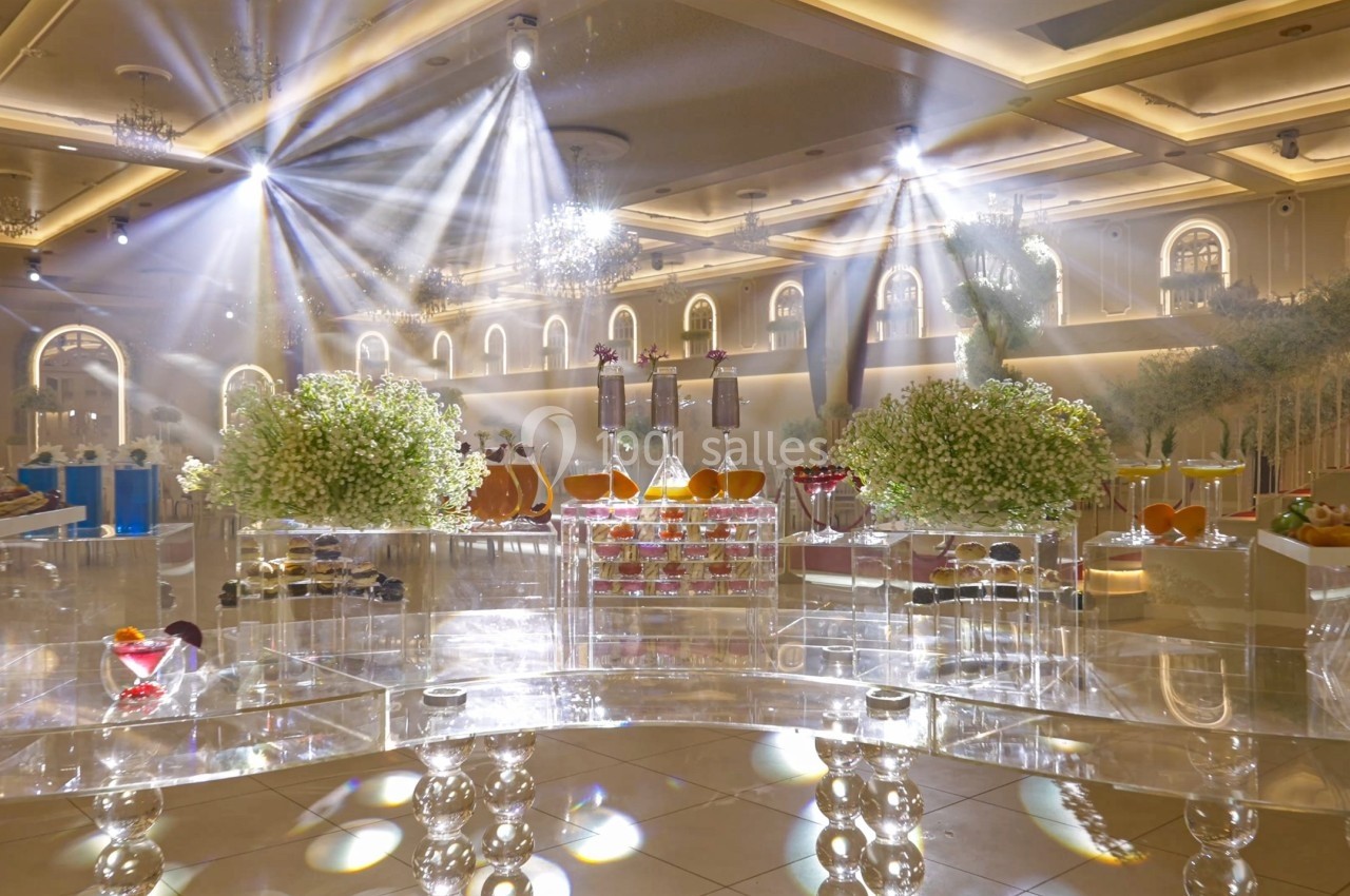 Salle de réception élégante avec éclairage lumineux, tables transparentes et décorations florales blanches.