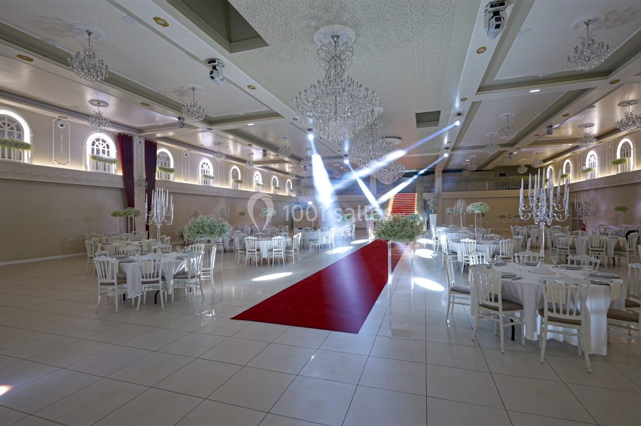 Salle de réception élégante avec tables dressées, lustres suspendus et tapis rouge central éclairé par des projecteurs.