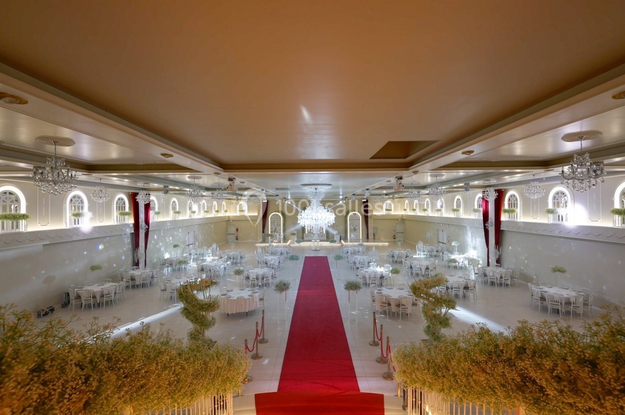Salle de réception élégante avec tapis rouge central, tables décorées et grand lustre au plafond.