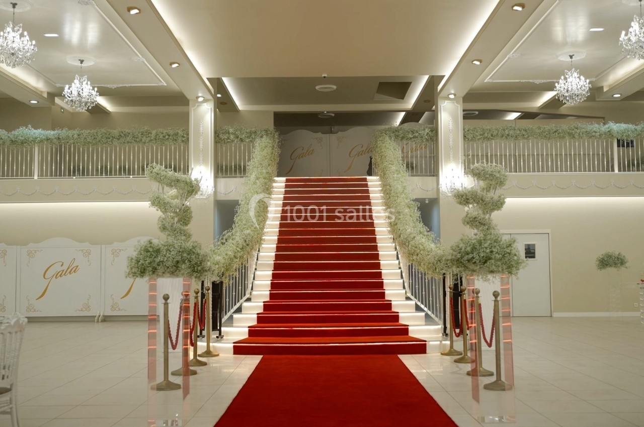 Escalier central avec tapis rouge, décoré de plantes et éclairé par des lustres dans une salle de réception élégante.