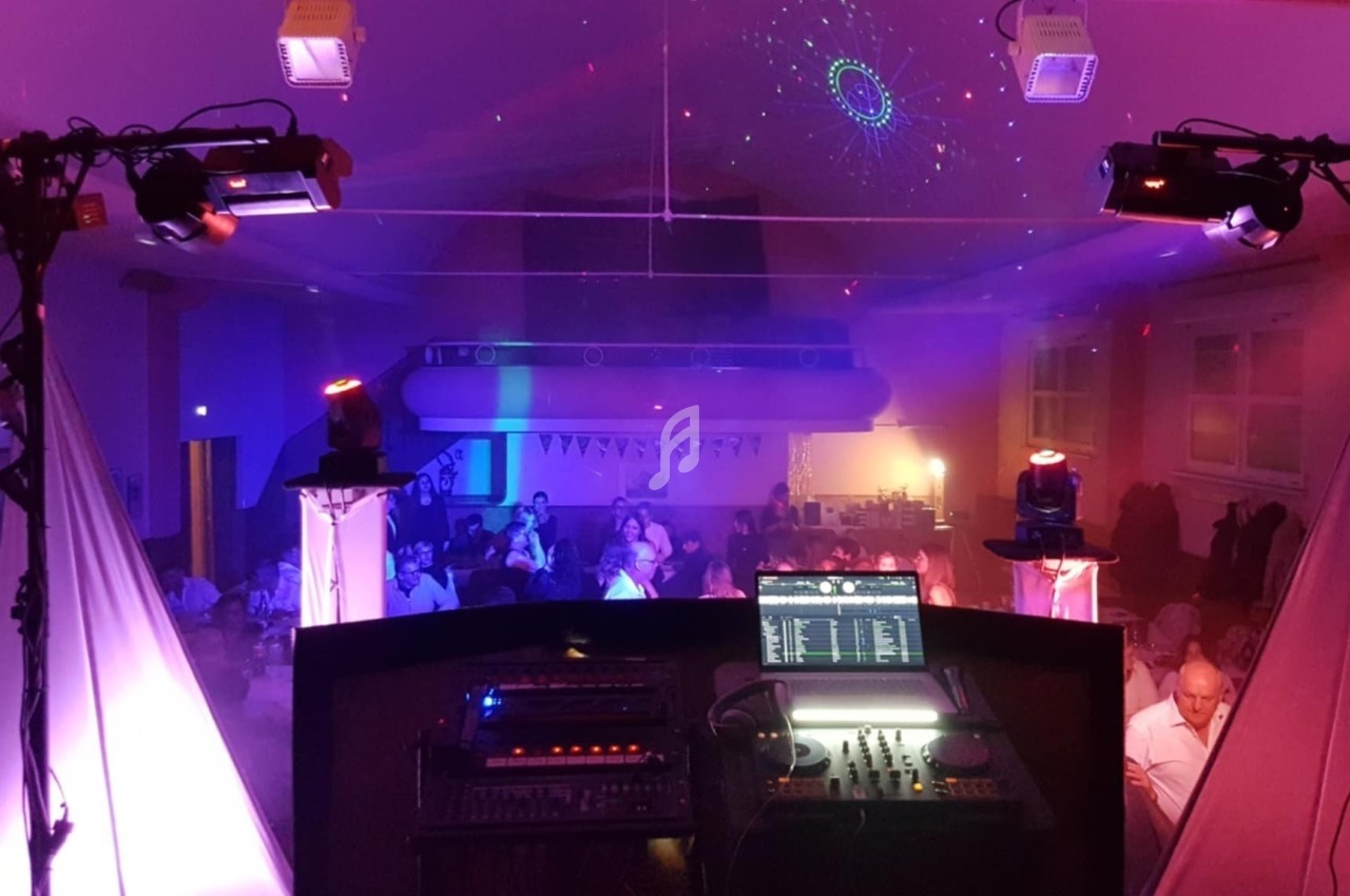 Vue depuis une cabine de DJ dans une salle éclairée par des jeux de lumière, avec des invités assis en arrière-plan.