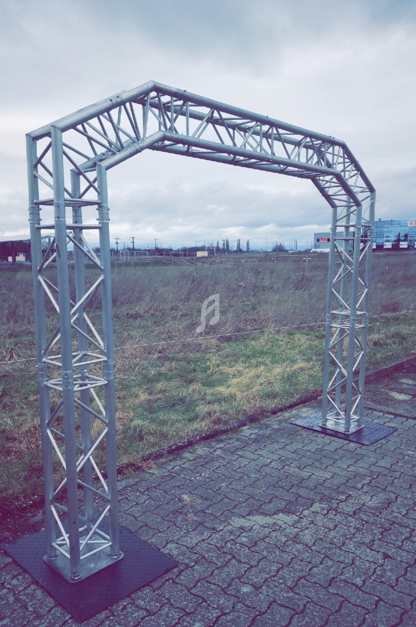 Structure métallique en forme d'arche installée sur un sol pavé, avec un paysage extérieur nuageux en arrière-plan.