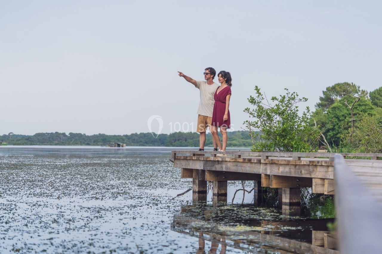 Un couple debout sur un ponton en bois observe un lac entouré de végétation.