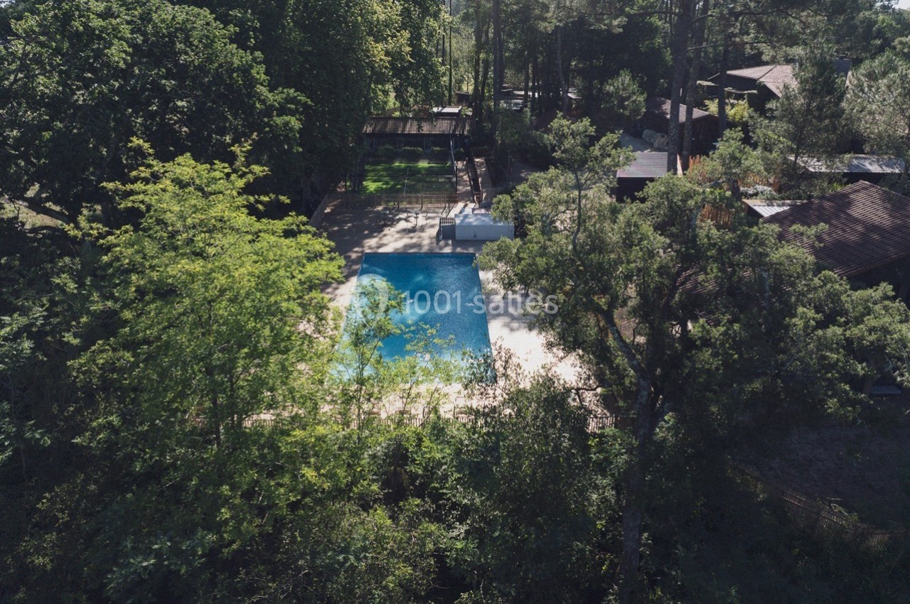 Vue aérienne d'une piscine entourée de végétation dense et de bâtiments en bois dans un cadre naturel.