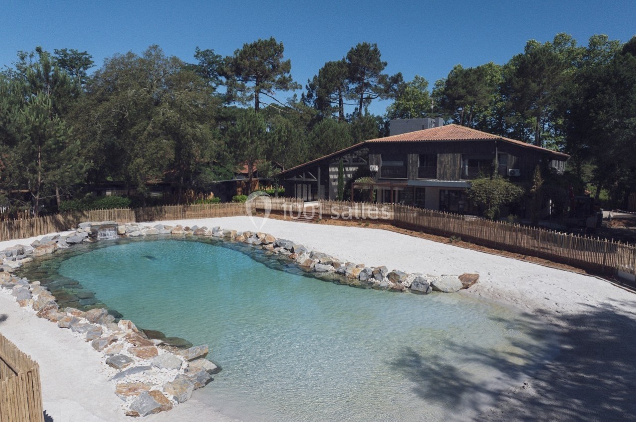 Maison en bois entourée d'arbres, avec un étang artificiel bordé de pierres et une plage de sable.