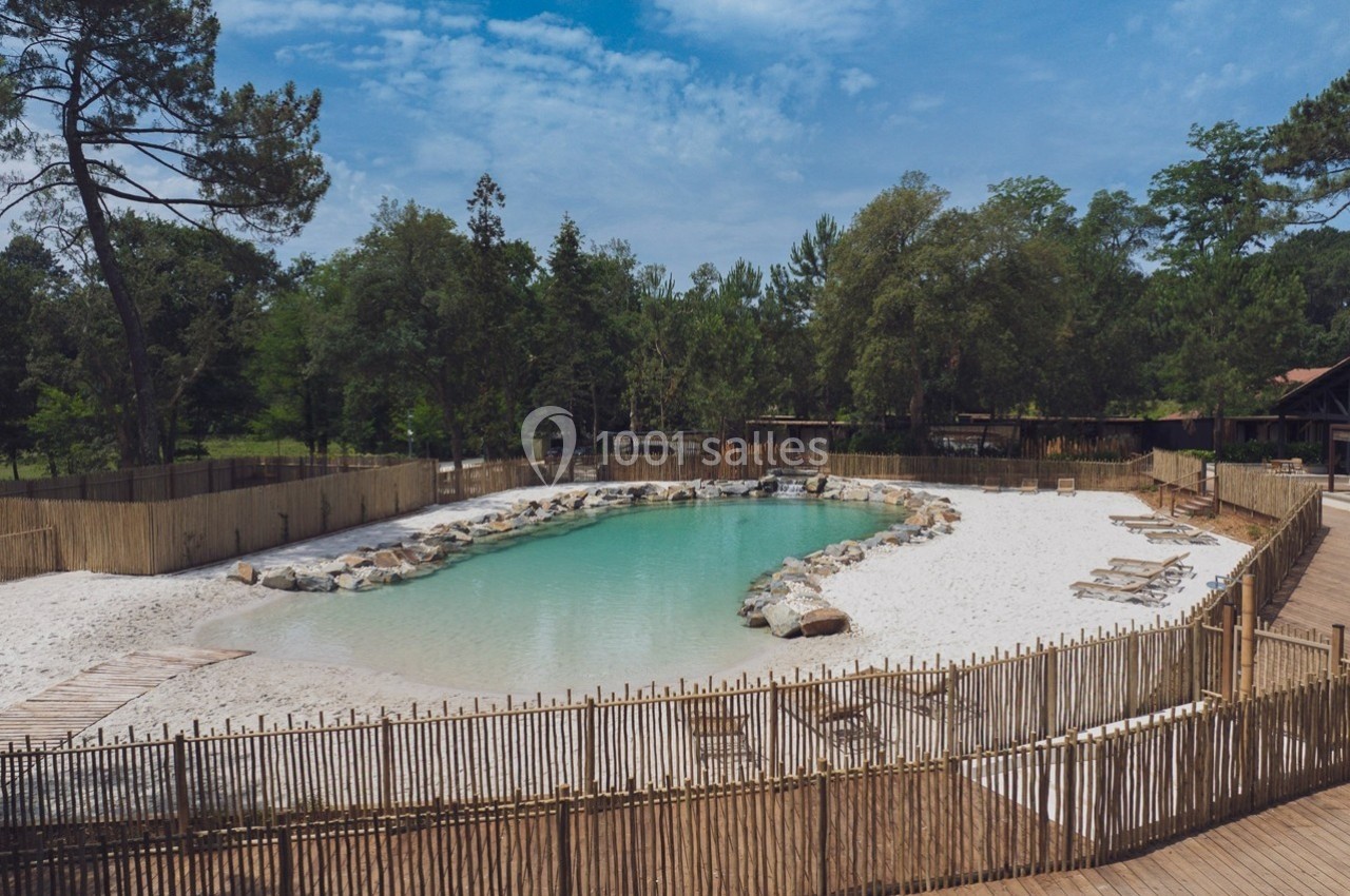 Piscine naturelle entourée de sable blanc, de rochers et de végétation, avec des transats en bois disposés autour.