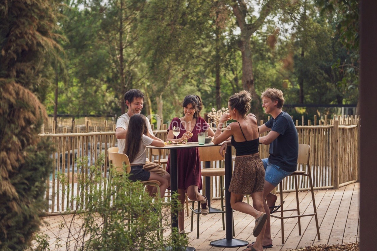 Des personnes discutent et partagent un repas autour d'une table haute en extérieur, entourées de verdure.