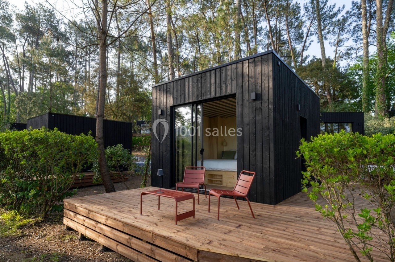 Cabane moderne en bois noir avec terrasse en bois, deux chaises rouges et table, entourée de végétation et de pins.