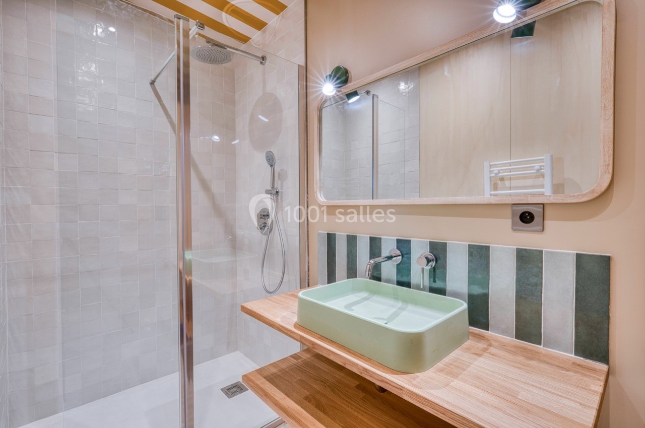 Salle de bain moderne avec douche vitrée, vasque verte sur un plan en bois et miroir rectangulaire.