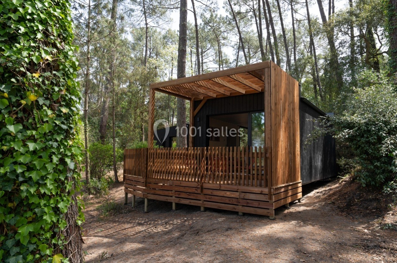 Cabane en bois moderne avec terrasse couverte, entourée de pins et de végétation dense en forêt.
