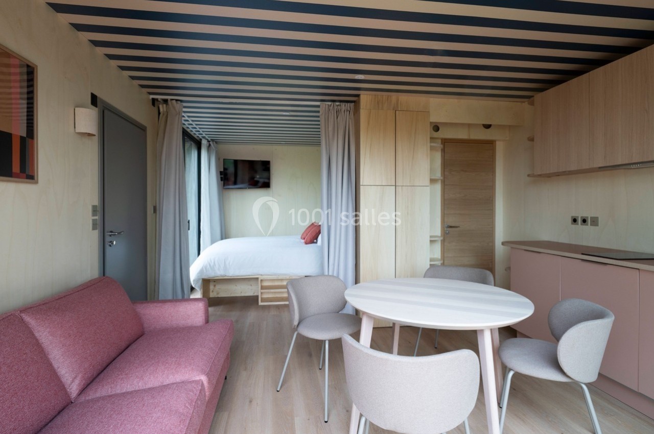 Intérieur d'un petit appartement moderne avec salon, table à manger, cuisine et lit visible en arrière-plan.