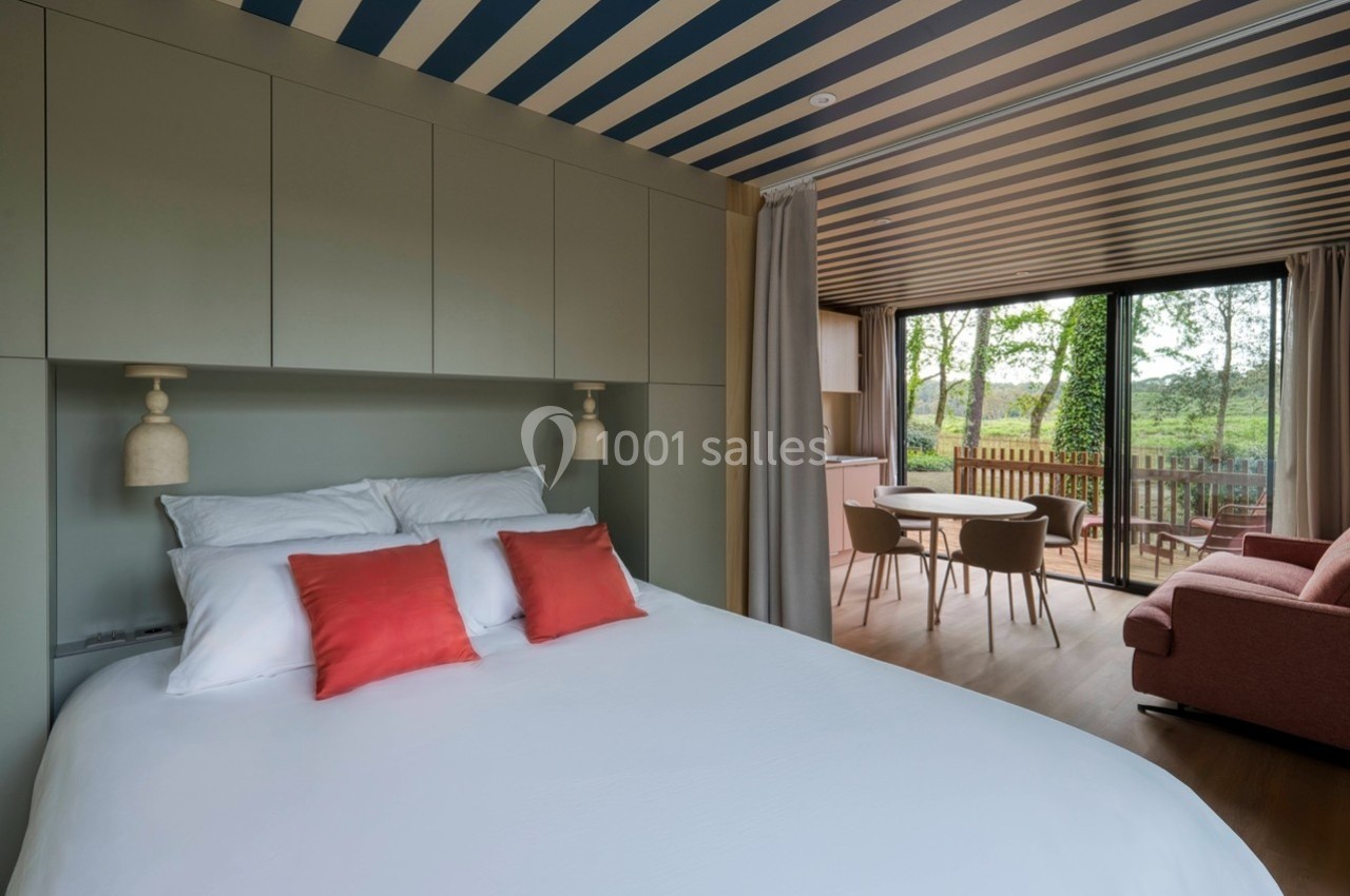 Chambre moderne avec lit double, coussins rouges, coin salon, table à manger et vue sur la nature à travers une baie vitrée.