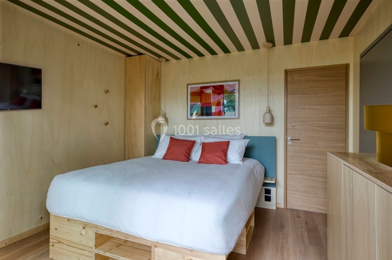 Chambre lumineuse avec lit double, murs en bois clair, plafond rayé vert et blanc, et décoration colorée.