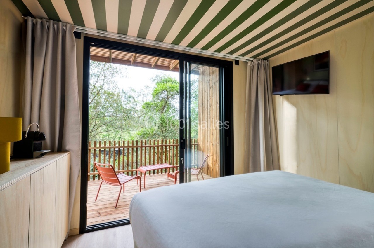 Chambre lumineuse avec lit, vue sur une terrasse en bois équipée de chaises, donnant sur un paysage arboré.